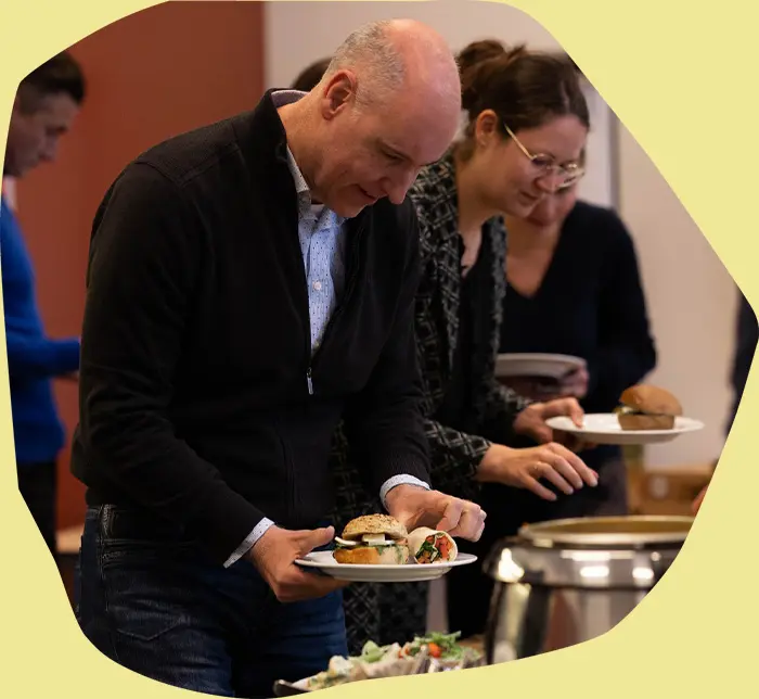Deelnemers van een training scheppen op bij het lunchbuffet; een man op de voorgrond houdt een bord met een belegd broodje en een wrap vast.
